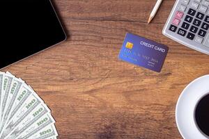 Top view of a blue credit card, tablet, US banknotes, pencil, calculator, and a white coffee cup on a wooden table in the office. Space for text. Wood texture background photo