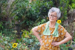 Portrait of an elderly woman exercises by gesturing raise both arms, smiling, and looking at the camera while standing in a garden. Space for text. Concept of aged people and healthcare photo