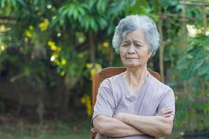 Senior woman with short gray hair arms crossed and looking at the camera while sitting on a wooden chair in the garden. Space for text. Concept of aged people and relaxation photo