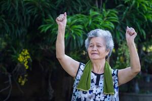 Senior woman wearing casual clothing, smiling, and exercise by gesturing raise both arms while standing in a garden. Concept of aged people and healthcare photo