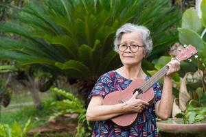 alegre un mayor mujer jugando el ukelele mientras en pie en un jardín. relajante por canto y jugar pequeño guitarra contento y disfrutar vida después saliente. concepto de Envejecido personas y cuidado de la salud foto