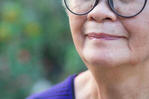 Close-up of half-face a senior woman wearing glasses, smiling and looking at the camera while standing in a garden. Space for text. Concept of aged people and healthcare photo