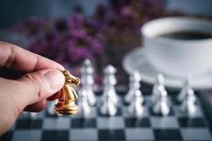 Close-up of hand holding a gold knight on a chessboard with a white coffee cup blur background. Business game competitive strategy. Concept of strategy for business photo