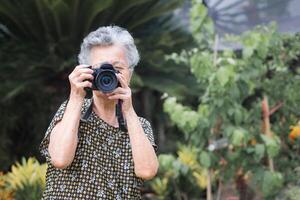 Senior woman with short gray hair shooting photos by a digital camera while standing outdoors. Space for text. Concept of aged people and photography