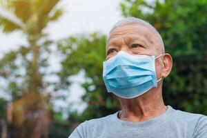 un retrato de un mayor hombre vistiendo un cara máscara mirando arriba mientras en pie en un jardín. máscara para proteger virus, COVID-19, coronavirus, bacterias, y más. concepto de Envejecido personas y cuidado de la salud foto