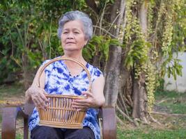 An elderly Asian woman holding a basket and looking at the camera, smiling while sitting on a chair in a garden. Space for text. Concept of aged people and relaxation photo