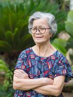 A portrait of an elderly woman with short gray hair wearing glasses, smiling with arms crossed, and looking at the camera while standing in a garden. Concept of aged people and healthcare photo