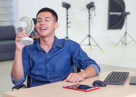 Asian young man's sitting at an office chair holding a small plastic fan blowing to his face because of hot weather. He's close-eyes and smiles with relaxation after using a fan. Studio workplace photo