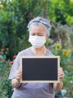 retrato de un mayor mujer vistiendo un cara máscara y participación un pizarra mientras en pie en un jardín. espacio para texto. concepto de Envejecido personas y cuidado de la salud foto