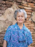 A portrait of an elderly Asian woman smiling and looking at the camera while standing with old brick wall background. Concept of aged people and relaxation photo