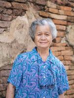 A portrait of an elderly Asian woman smiling and looking at the camera while standing with old brick wall background. Concept of aged people and relaxation photo