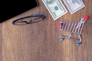 Top view of a stack of one hundred dollars and fifty dollars, glasses, mini shopping cart, and tablet on wooden desk in the office. Space for text. Wood texture background. E-commerce concept photo