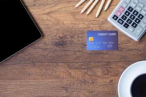 Top view of a blue credit card, pencil, calculator, coffee cup, and tablet on wooden table in the office. Space for text. Wood texture background. Business and finance concept photo