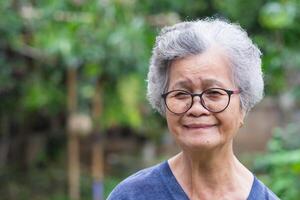 A senior woman with short gray hair wearing glasses smiling and looking at the camera while standing in a garden. Space for text. Concept of aged people and healthcare photo