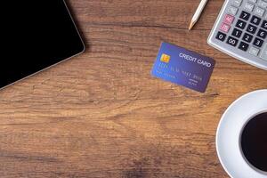 Top view of a blue credit card, pencil, calculator, coffee cup, and tablet on wooden table in the office. Space for text. Wood texture background. Business and finance concept photo