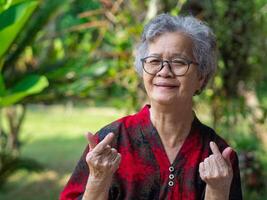 retrato de felicidad mayor mujer sonriente y manos haciendo mini corazón mientras en pie en un jardín. concepto de Envejecido personas y cuidado de la salud foto