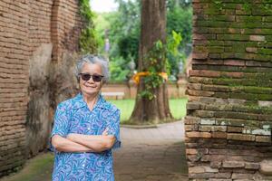 Senior woman with short gray hair wearing sunglasses, arms crossed, smiling, and looking at the camera while standing with old brick wall background. Concept of aged people and relaxation photo