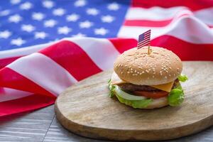 The close-up image of the homemade hamburger with lettuce and cheese has an American flag pin on the hamburger placed on a wooden chopping board with an American flag background on a wooden table photo