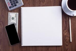 Top view of smartphone, sketchbook, pencil, calculator, US dollars, and a white coffee cup on wooden table. Template for text. Wood texture background. Concept of drawing and technology photo