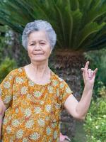 Senior woman with short gray hair showing fingers I love you a symbol, smiling and looking at the camera while standing in a garden. Concept of aged people and healthcare photo