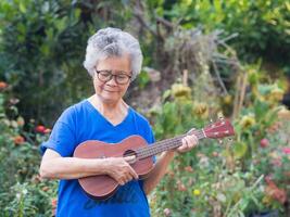 retrato de mayor mujer jugando ukelele en su jardín. relajante por canto y jugar pequeño guitarra contento y disfrutar vida después jubilado. concepto de antiguo personas y salud cuidado. espacio para texto foto