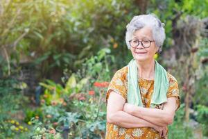 Portrait of an elderly woman with short gray hair wearing glasses, arms crossed, smiling, and looking at the camera while standing in a garden. Concept of age people and healthcare photo