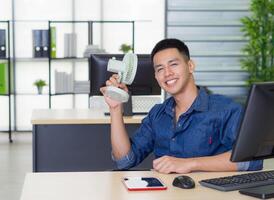 Asian young man's sitting at an office chair holding a small plastic fan blowing to his face because of hot weather. He's looking at the camera and smiles with relaxation the studio workplace photo