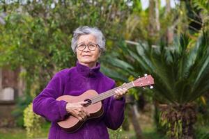 Portrait of a senior woman with short gray hair playing the ukulele, smiling, and looking up while standing in a garden. Concept of aged people and relaxation photo