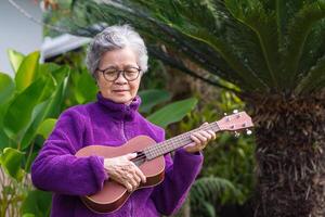 Portrait of an elderly Asian woman with short gray hair playing the ukulele while standing in a garden. Concept of aged people and relaxation photo