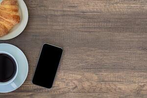 Top view of a table with a smartphone, a white coffee cup, and croissants placed on a wooden table. Work from home. Space for text photo