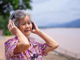 Portrait of a happy elderly woman wearing wireless headphones listening to music while standing side the river. Space for text. Concept of aged people and relaxation photo