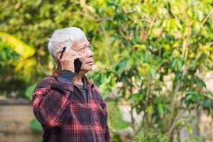 Elderly man using a smartphone and looking up while standing in a garden. Space for text. Concept of aged people and communication photo