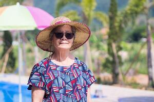 A portrait of an elderly woman wearing sunglasses and straw hat while standing side swimming pool. Space for text. Concept of old people and holiday photo