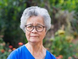 A portrait of an elderly Asian woman with short gray hair, wearing glasses, smiling, and looking at the camera while standing in a garden. Space for text. Concept of aged people and healthcare photo