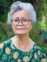 Portrait of a senior woman with short gray hair, wearing glasses, smiling, and looking at the camera while standing in a garden. Space for text. Concept of aged people and healthcare photo