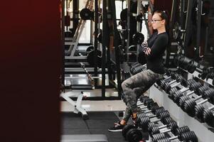 hermosa niña con manos en pesas mirando en su reflexión en el espejo en un Deportes salón. Delgado joven mujer es tomando pesas en frente de un espejo en aptitud club foto