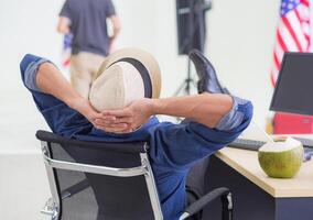 Back view of a young man wearing a hat and placing both hands on the back of the neck while sitting on a chair in the office. Holiday coming soon. Holiday concept photo