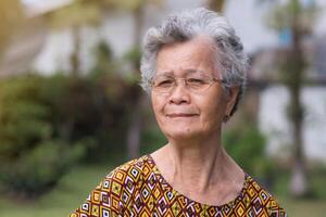 Portrait of an elderly Asian woman with short gray hair, wearing glasses, smiling and looking at the camera while standing in a garden. Space for text. Concept of aged people and healthcare photo
