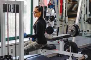 sonriente hermosa niña con pesado pesas en frente de el espejo en un Deportes salón. fuerte joven hermosa mujer es comprometido en gimnasio con pesas en frente de un espejo foto