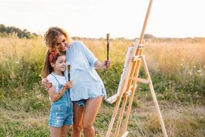 Young attractive mother teaches daughter painting in summer park. Outdoors activity for school age children concept. photo