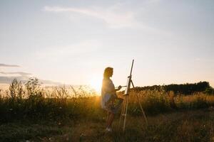 silueta de un rubia niña artista. dama pinturas un pintura en el lona con el ayuda de pinturas un de madera caballete mantiene el fotografía. verano es un soleado día, puesta de sol. foto