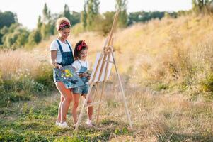 Young attractive mother teaches daughter painting in summer park. Outdoors activity for school age children concept. photo