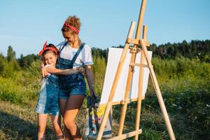 Beauty mother paint with her little daughter. Stylish woman drawing the picture with little girl. Cute kid in a white t-shirt and blue jeans. Mother's Day. photo