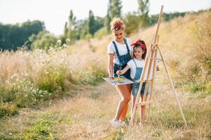 Young attractive mother teaches daughter painting in summer park. Outdoors activity for school age children concept. photo