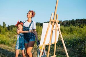 Beauty mother paint with her little daughter. Stylish woman drawing the picture with little girl. Cute kid in a white t-shirt and blue jeans. Mother's Day. photo