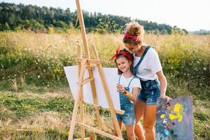 belleza madre pintar con su pequeño hija. elegante mujer dibujo el imagen con pequeño muchacha. linda niño en un blanco camiseta y azul vaqueros. de la madre día. foto