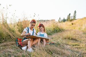 belleza madre pintar con su pequeño hija. elegante mujer dibujo el imagen con pequeño muchacha. linda niño en un blanco camiseta y azul vaqueros. de la madre día. foto