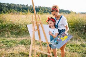 belleza madre pintar con su pequeño hija. elegante mujer dibujo el imagen con pequeño muchacha. linda niño en un blanco camiseta y azul vaqueros. de la madre día. foto