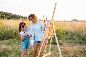 Beauty mother paint with her little daughter. Stylish woman drawing the picture with little girl. Cute kid in a white t-shirt and blue jeans. Mother's Day. photo