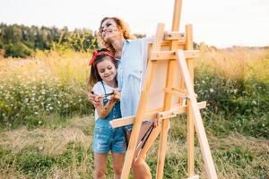 joven atractivo madre enseña hija pintura en verano parque. al aire libre actividad para colegio años niños concepto. foto